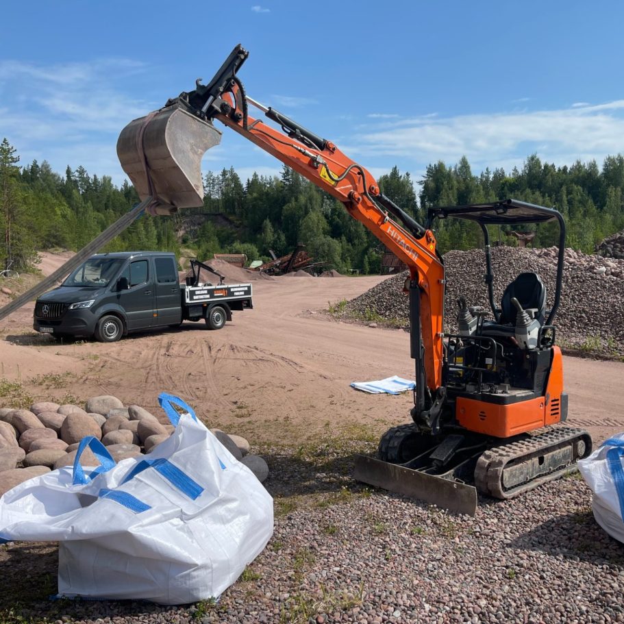 Verladung der Findlinge in BigBag Kleiner Bagger mit hochgestelltem Ausleger auf Baustelle mit Lkw und Schotter.
