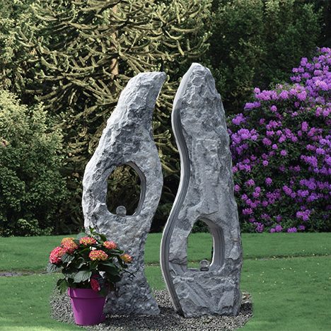 Zwei stilisierte Steinskulpturen neben einem blühenden Topf mit bunten Blumen.